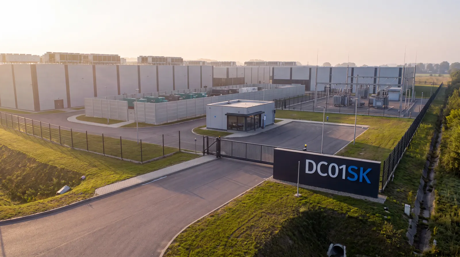 Concept visualisation of a build-to-suit data center campus on the DC01SK site: an access road leading to a gated entrance and gatehouse, with long halls set behind the perimeter fence.