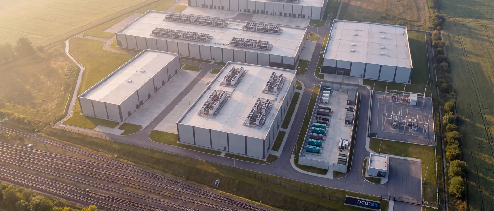 Aerial concept visualisation of a build-to-suit data center campus on the DC01SK site: several long halls with rooftop cooling plant, linked by internal service roads.